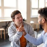 Young happy man with Down syndrome with his mentoring friend celebrating success indoors at school.